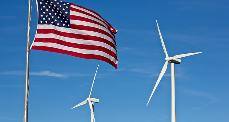 An american flag flies in the foreground, with two wind power generator windmills turn in the background