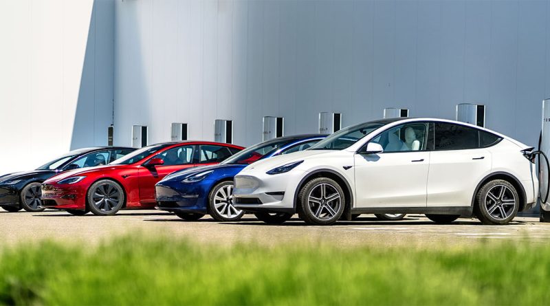 Multiple Tesla electric vehicles including Model 3 and Model Y charging at Tesla Supercharger stations outside a modern factory facility