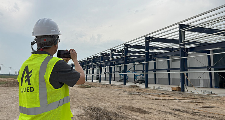 a construction worker, wearing a hard hat and high-visibility vest with "ALLIED" written on it, taking a photo or video of a building under construction