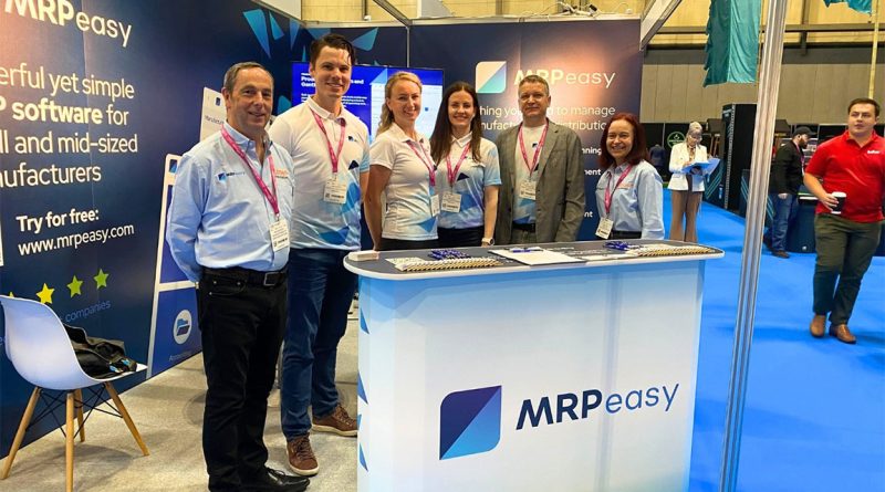 Five MRPeasy team members standing behind a branded booth at a trade show, smiling at the camera. The booth displays banners promoting MRP software for small and mid-sized manufacturers, with brochures and pens arranged on the counter. The event space features blue flooring and other exhibitors in the background