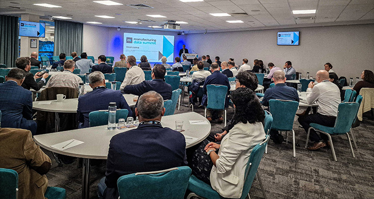 a conference room with an audience seated at circular tables, facing a large projection screen at the front of the room