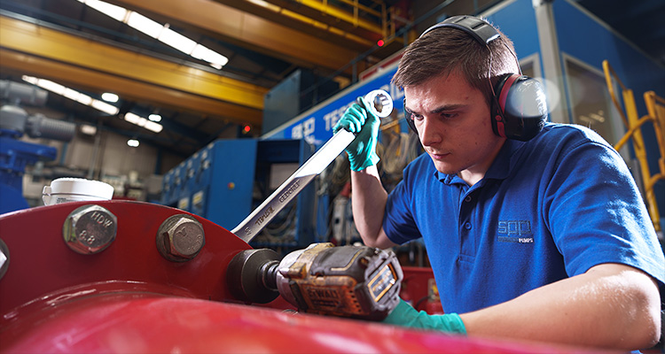 a person in a factory setting wearing a blue polo shirt, green gloves, and ear defenders while using a power tool on a piece of machinery