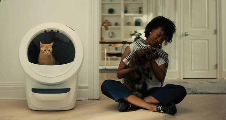 a woman sitting on the floor with a dark-colored cat in her lap. Next to her is a white, automated self-cleaning litter box, the Litter-Robot 4 by Whisker. An orange cat is visible inside the litter box