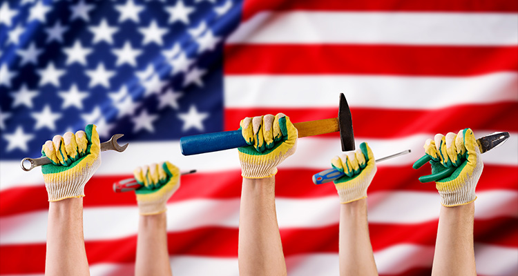 several hands, clad in yellow and green work gloves, each holding different hand tools such as a wrench, screwdrivers, a hammer, and a utility knife or chisel. These hands are raised in the foreground against a backdrop of a large, blurred American flag