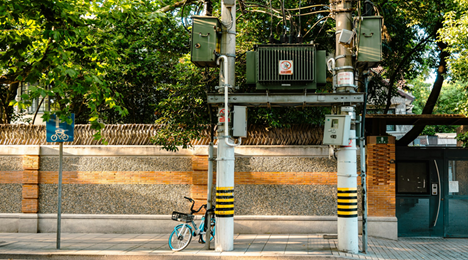 Industrial electrical transformer mounted on two white poles with black and yellow hazard stripes, overhead cables and green utility boxes, a blue bicycle parked beneath it on a sidewalk beside a brick and stone wall with trees in the background.