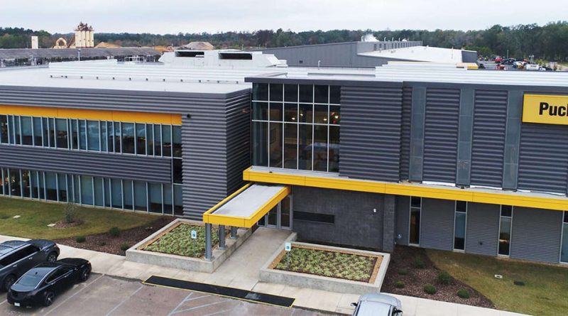 Modern Puckett CAT dealership building with gray metal exterior, large glass windows and yellow CAT signage, viewed from above with parked vehicles in front.